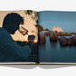 The open book "Wine & Travel Mediterranean Islands" by Assouline shows a man examining Sicilian wine on the left page and a dusk coastal scene with rocks and a building on the right.