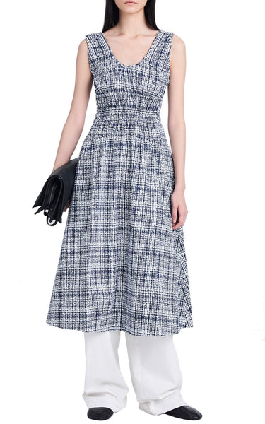 Woman modeling a Proenza Schouler Penny Grid Poplin Dress with a smocked bodice and a blue and white pattern, paired with white wide-legged pants and holding a black clutch.