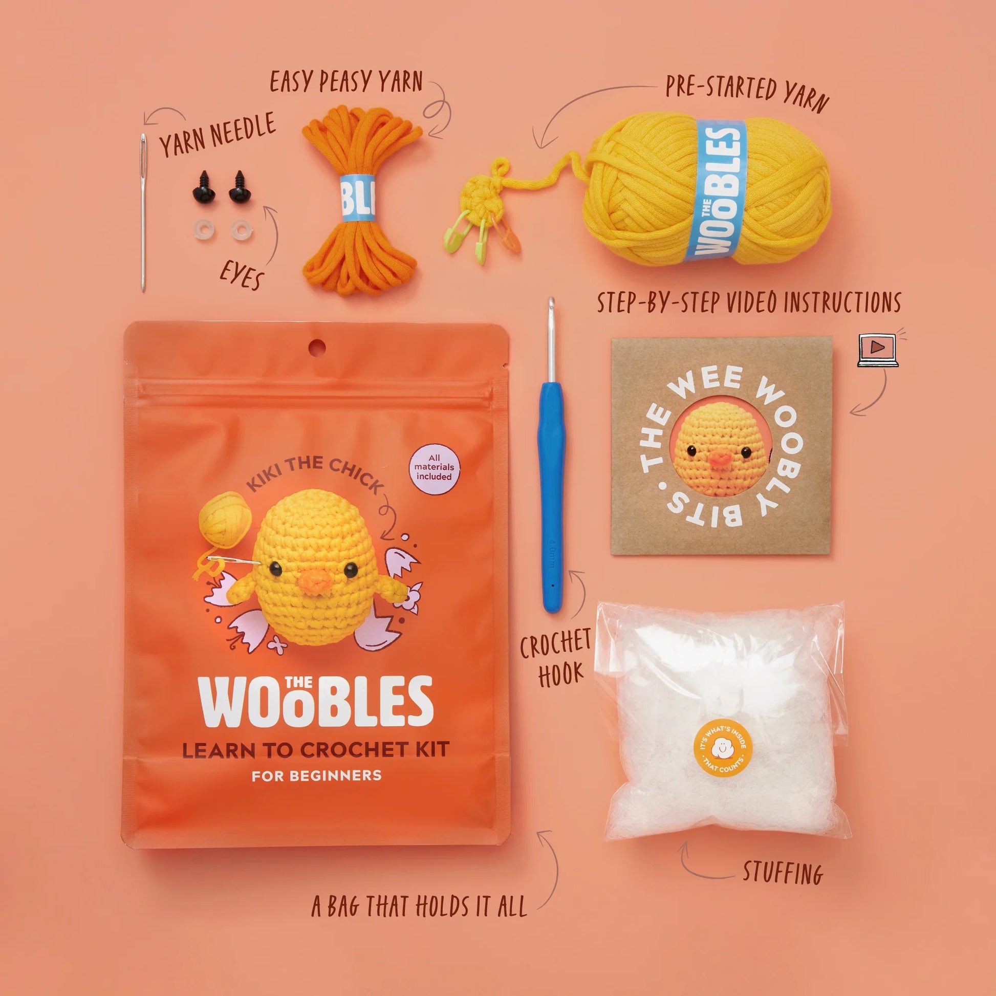 Flat lay of The Woobles Kiki the Chick Beginner Crochet Kit, featuring labeled items—including yarn, hook, needle, eyes, stuffing, instructions, and a resealable bag—on a peach background.