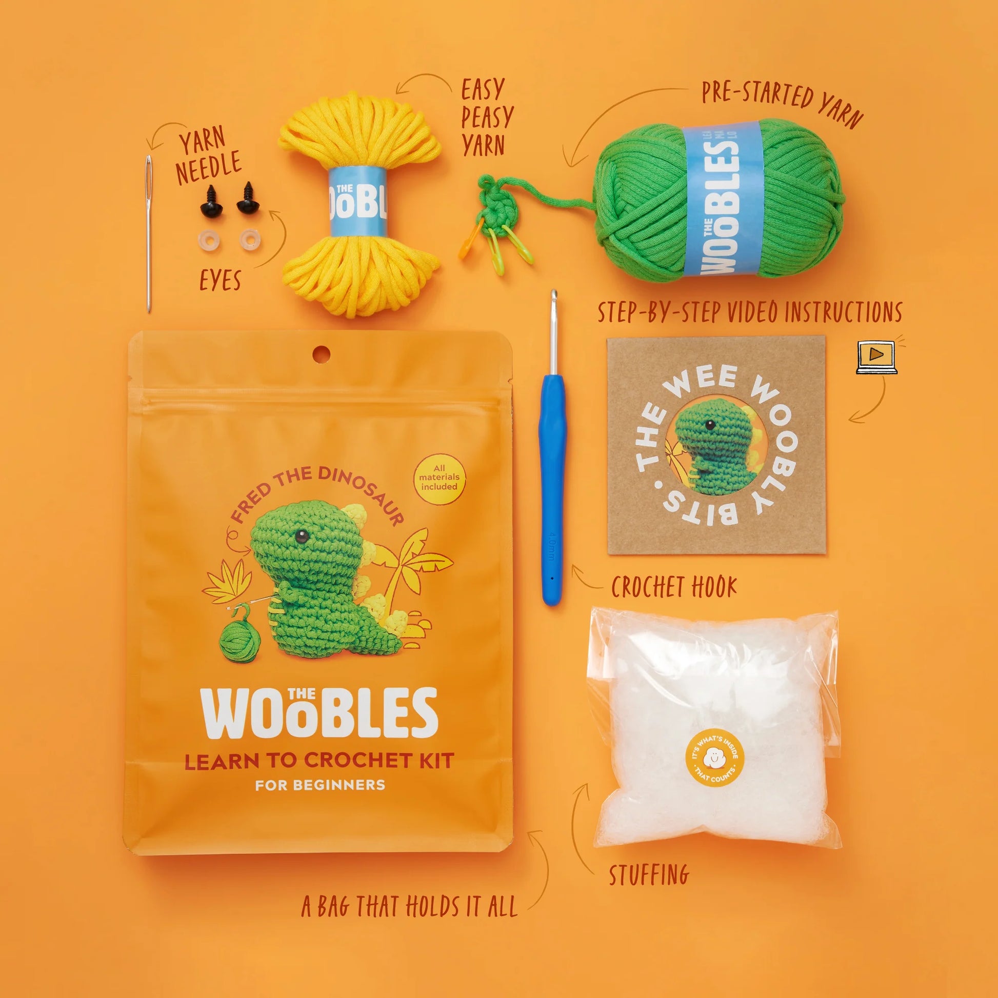 The Woobles Fred the Dinosaur Beginner Crochet Kit includes everything you need to make your own Fred the Dino—yarn, hook, yarn needle, safety eyes, stuffing, simple step-by-step instructions (including how to start with a magic loop), and a convenient storage bag—all presented on an eye-catching orange background.