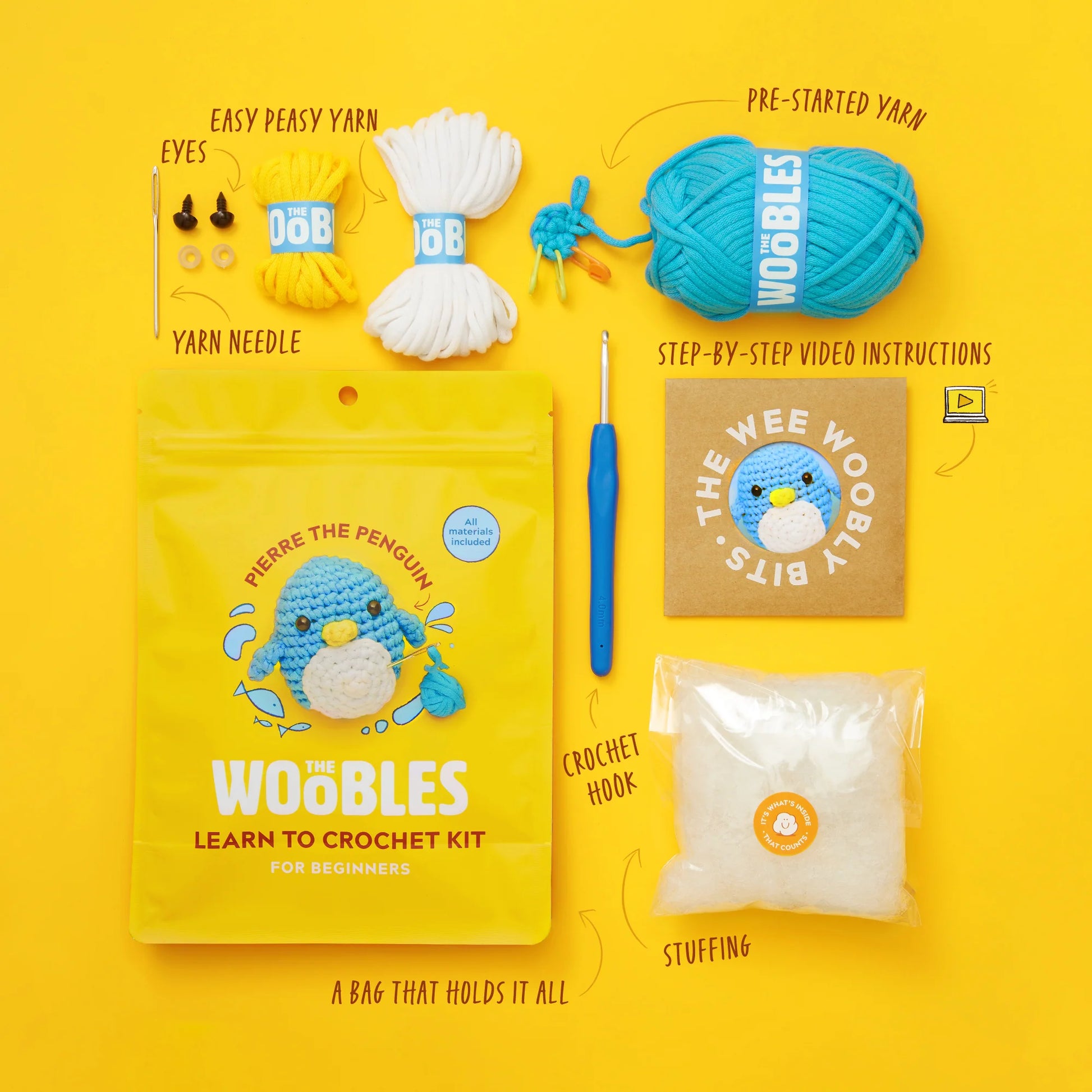 Pierre the Penguin Beginner Crochet Kit by The Woobles displayed on a yellow background, featuring yarn, safety eyes, needle, crochet hook, stuffing, instructions, and packaging with the adorable blue penguin Pierre. Ideal for anyone interested in trying beginner crochet or discovering the joy of a Woobles penguin kit.