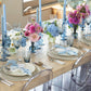 A dining table set with light blue and white decor, pastel flowers in vases, Kim Seybert Iris Tall Candle Holder Set, hand-blown glassware, and transparent chairs.