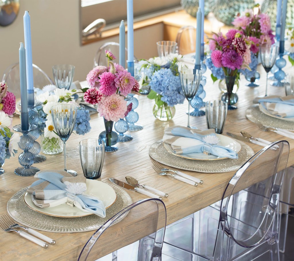 A dining table set with light blue and white decor, pastel flowers in vases, Kim Seybert Iris Tall Candle Holder Set, hand-blown glassware, and transparent chairs.