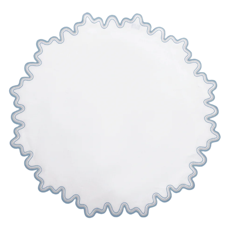 The Matouk Wave Tabletop Collection in Hazy Blue features a round white linen tabletop with a wavy blue embroidered edge and is OEKO-TEX certified for safety and style.