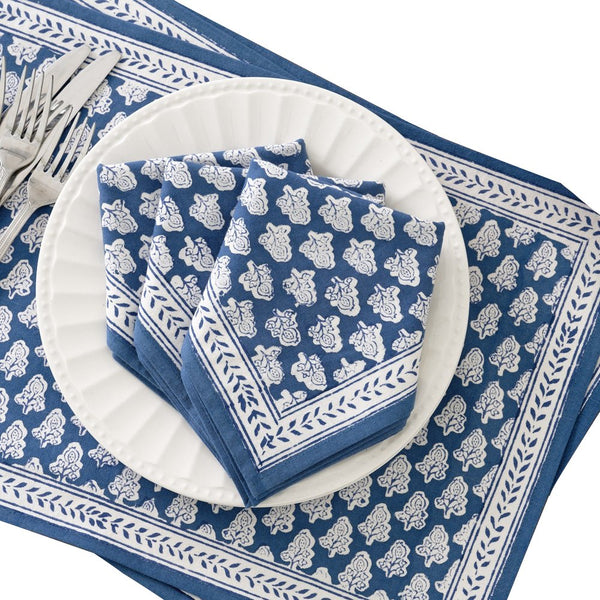 A white plate sits on a Pomegranate Inc. Pom Buti Denim Collection floral blue and white placemat with three folded matching napkins on top. On the left, there are three forks and a knife, setting the stage for an elegant dining experience.