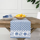 A wooden table with a blue and white patterned table runner from the Pomegranate Inc Pom Buti Denim Collection by Pomegranate Inc., featuring a stack of plates, silverware wrapped in napkins, and a bowl of lemons for an inviting dining experience.
