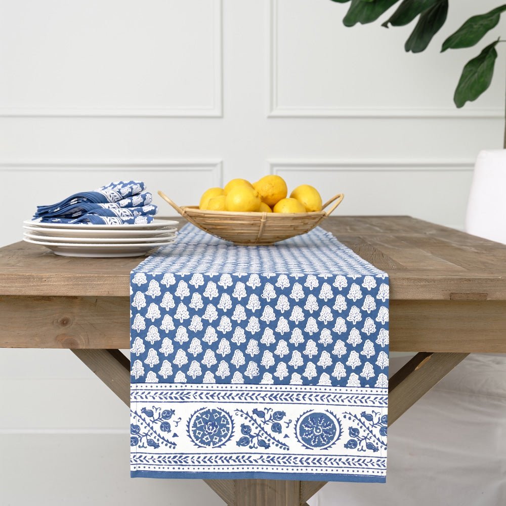 A wooden table with a blue and white patterned table runner from the Pomegranate Inc Pom Buti Denim Collection by Pomegranate Inc., featuring a stack of plates, silverware wrapped in napkins, and a bowl of lemons for an inviting dining experience.