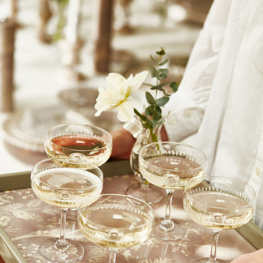 A person in a white shirt holds an Addison Ross Medium Chinoiserie Tray Collection piece with five champagne coupes and a small vase featuring a white flower and greenery.