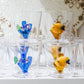 Four glass tumblers from Ichendorf Milano’s Marine Garden Drinkware Collection—two featuring blue and purple fish, two with yellow—are artfully displayed on a white shelf.