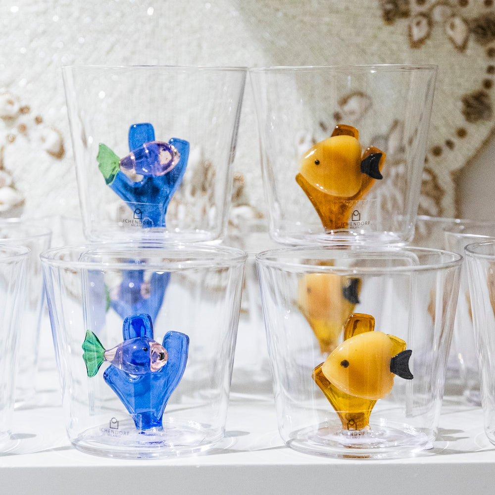 Four glass tumblers from Ichendorf Milano’s Marine Garden Drinkware Collection—two featuring blue and purple fish, two with yellow—are artfully displayed on a white shelf.