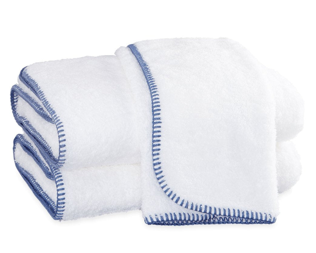 A stack of Matouk Whipstitch Bath Collection towels, featuring white fabric and blue terry whipstitching, is neatly arranged on a white surface.