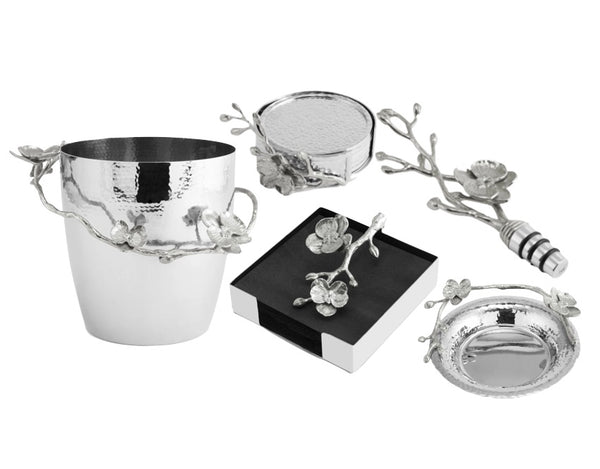 The Michael Aram White Orchid Barware collection features a silver ice bucket, coasters, bottle stopper, and napkin holder with elegant floral motifs, beautifully arranged on a white background to highlight their purity.