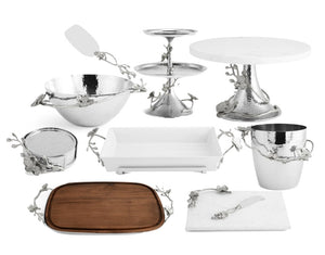 The Michael Aram White Orchid Serveware Collection features metal, wood, and ceramic serving pieces with metallic branch and leaf accents, beautifully arranged on a pure white background.