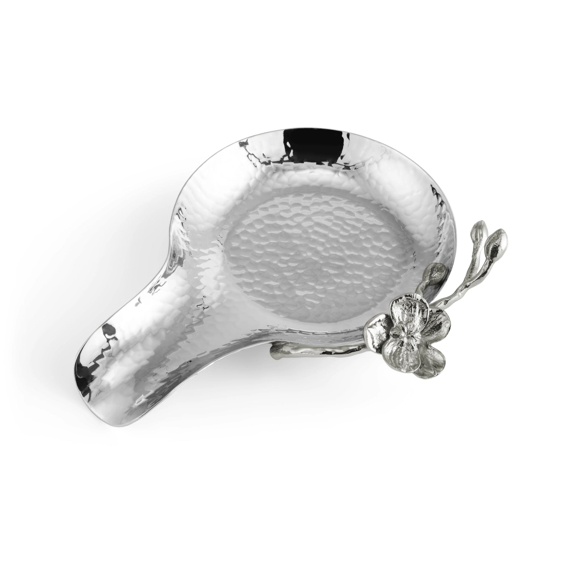 The Michael Aram White Orchid Serveware Collection includes a shiny, hammered silver spoon rest with a flower and branch detail on one side, evoking purity and elegance. Shown on a white background.