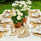 An outdoor table set for dining features white napkins, wine glasses, golden votive holders, and potted white flowers on a cream tablecloth with the Kim Seybert Java Table Runner as a centerpiece accent.