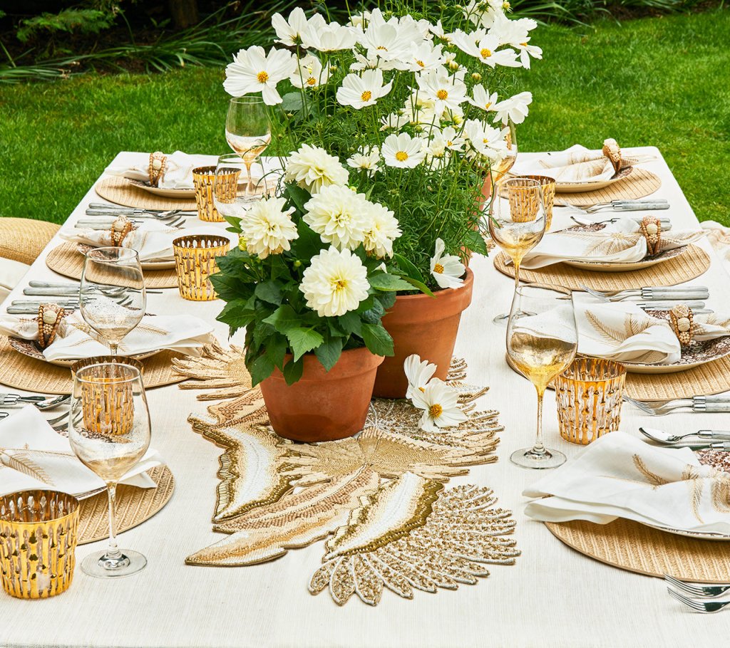 An outdoor table set for dining features white napkins, wine glasses, golden votive holders, and potted white flowers on a cream tablecloth with the Kim Seybert Java Table Runner as a centerpiece accent.