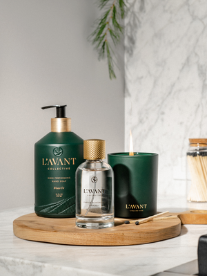 A bottle of L'Avant Collective Winter Fir Room Spray sits on a round wooden tray with holiday decor, enhancing comfort on a marble countertop next to a jar of matches.