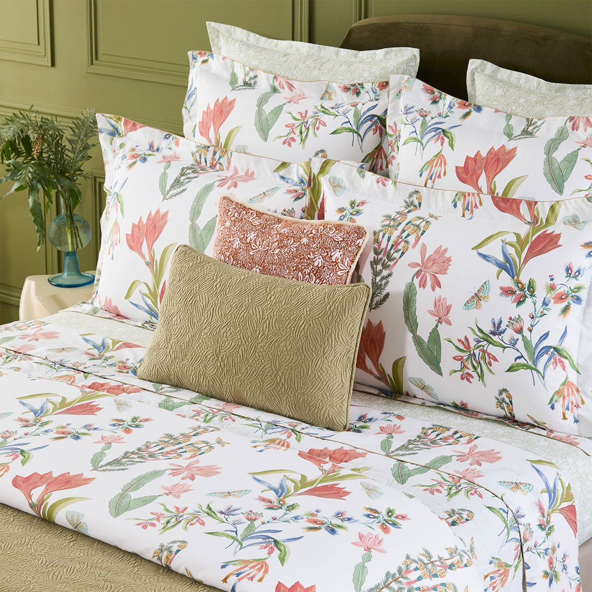 A neatly made bed with Yves Delorme's Eze Bedding Collection, featuring white organic cotton linen with a colorful floral design, and topped with two gold and peach decorative pillows.