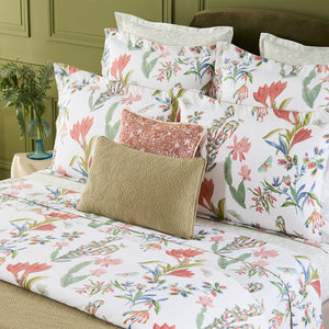 A neatly made bed with Yves Delorme's Eze Bedding Collection, featuring white organic cotton linen with a colorful floral design, and topped with two gold and peach decorative pillows.