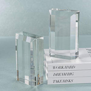 The Zodax Claire Crystal Glass Bookend Set holds and flanks three white books titled "WORK HARD," "DREAM BIG," and "TAKE RISKS" for elegant bookshelf decor on a light blue surface.