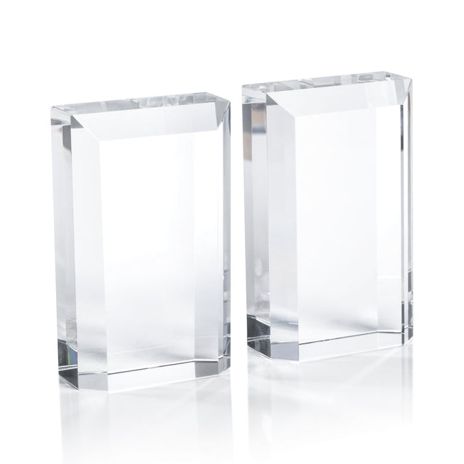The Zodax Claire Crystal Glass Bookend Set features two clear rectangular glass blocks with beveled edges, standing upright on a white surface for an elegant, minimalist display.