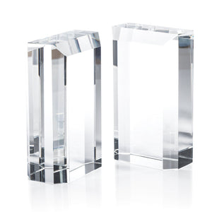The Claire Crystal Glass Bookend Set by Zodax features two clear rectangular glass blocks with sharp edges and clean lines, serving as elegant minimalist bookends and a sophisticated addition to any bookshelf decor.