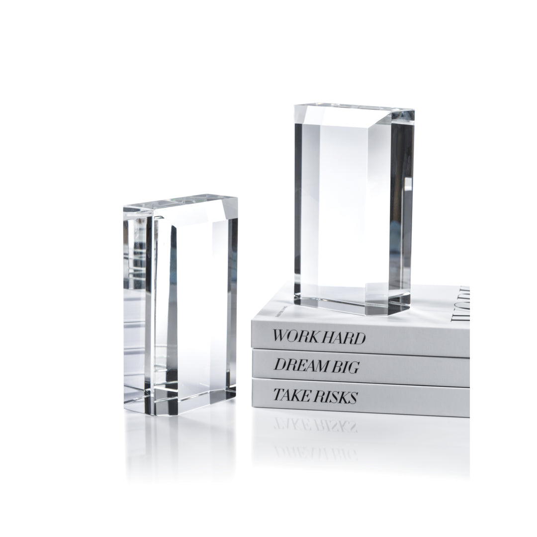 The Zodax Claire Crystal Glass Bookend Set features two clear rectangular bookends on a white surface, paired with books labeled "WORK HARD," "DREAM BIG," and "TAKE RISKS" for a modern, elegant touch to any space.
