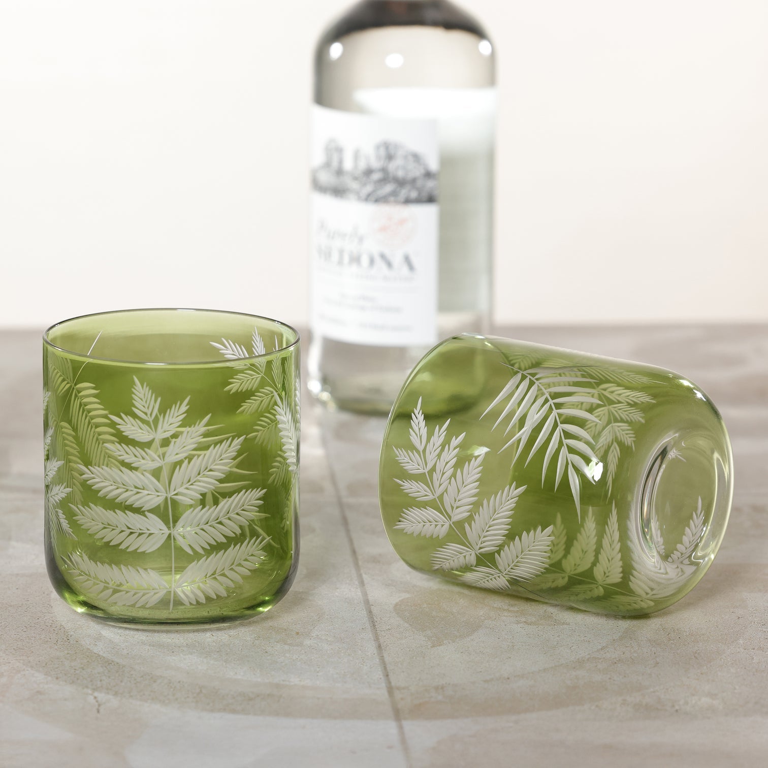The Zodax Barware Fern Leaf Rock Glass is shown in green, featuring an etched fern design—one upright, one on its side—with a clear bottle in the background on a stone surface, perfect for elevating your home bar.