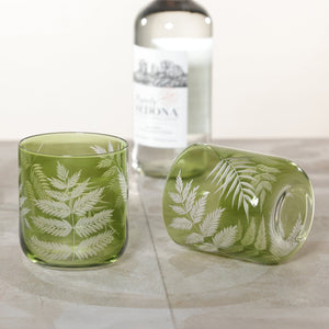 The Zodax Barware Fern Leaf Rock Glass is shown in green, featuring an etched fern design—one upright, one on its side—with a clear bottle in the background on a stone surface, perfect for elevating your home bar.