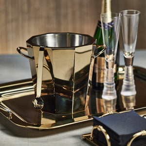 The Zodax Alessia Gold Double Wall Ice Bucket with matching scoop rests on a tray with two champagne flutes, a champagne bottle, and a stack of dark napkins.