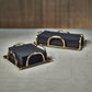 Two stacks of black square napkins rest in Zodax Alessia Gold Napkin Holders on a light surface, with a blurred brown background.