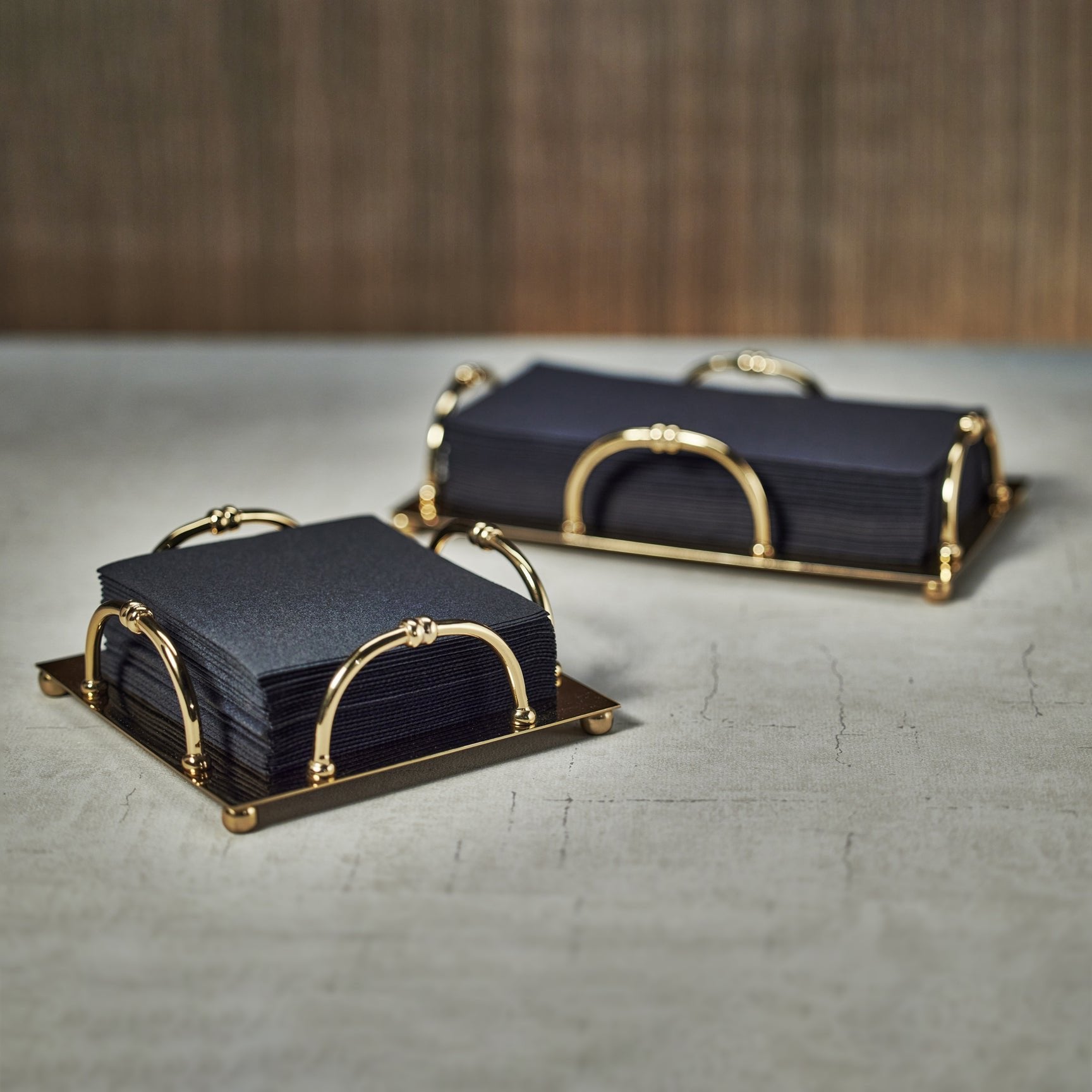 Two stacks of black square napkins rest in Zodax Alessia Gold Napkin Holders on a light surface, with a blurred brown background.