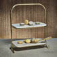 The Zodax Madeleine Marble Two-Tier Stand displays assorted macarons, with a gold serving tong on the lower tier—ideal for stylish entertaining. Set against a neutral brown background.