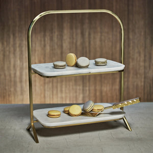 The Zodax Madeleine Marble Two-Tier Stand displays assorted macarons, with a gold serving tong on the lower tier—ideal for stylish entertaining. Set against a neutral brown background.
