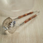 The Colony Bamboo and Nickel Serving Sets by Zodax, featuring shell-shaped tongs with bamboo handles, rest on a textured beige surface.