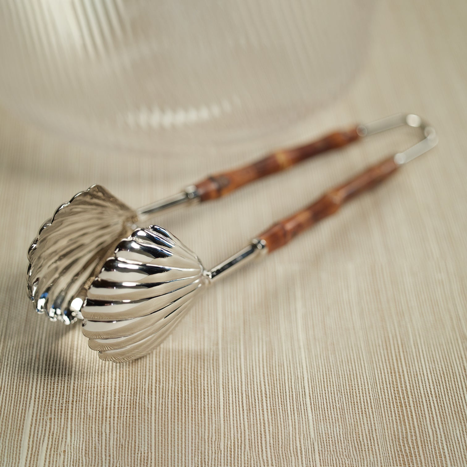 The Colony Bamboo and Nickel Serving Sets by Zodax, featuring shell-shaped tongs with bamboo handles, rest on a textured beige surface.