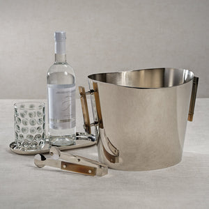 On a light tablecloth, The Berkeley Ice Tong with Horn Trim by Zodax is arranged alongside a metal ice bucket, a dotted glass on a tray, and a bottle of clear liquid.