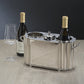 The Zodax Alsace Brass Two Bottle Wine Cooler in Polished Nickel holds two bottles of wine and a glass, displayed on a beige surface with a dark background.