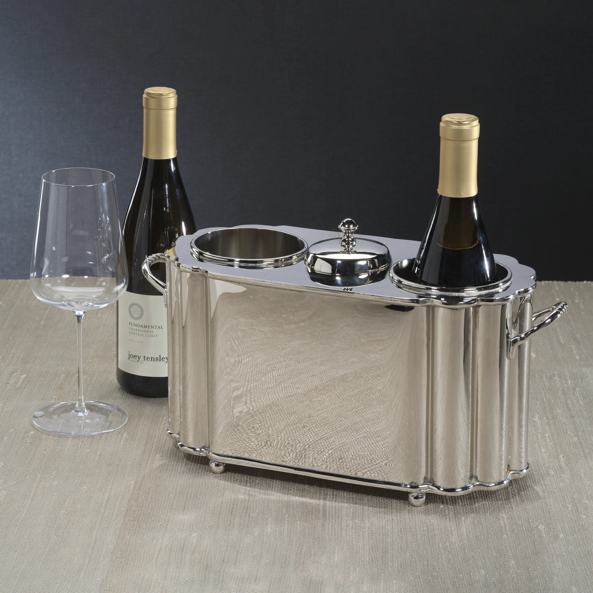The Zodax Alsace Brass Two Bottle Wine Cooler in Polished Nickel holds two bottles of wine and a glass, displayed on a beige surface with a dark background.