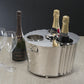 The Zodax Alsace Three Bottle Wine / Champagne Cooler in polished nickel holds bottles and ice, with a bottle of Krug champagne and two empty glasses beside it on a textured surface.