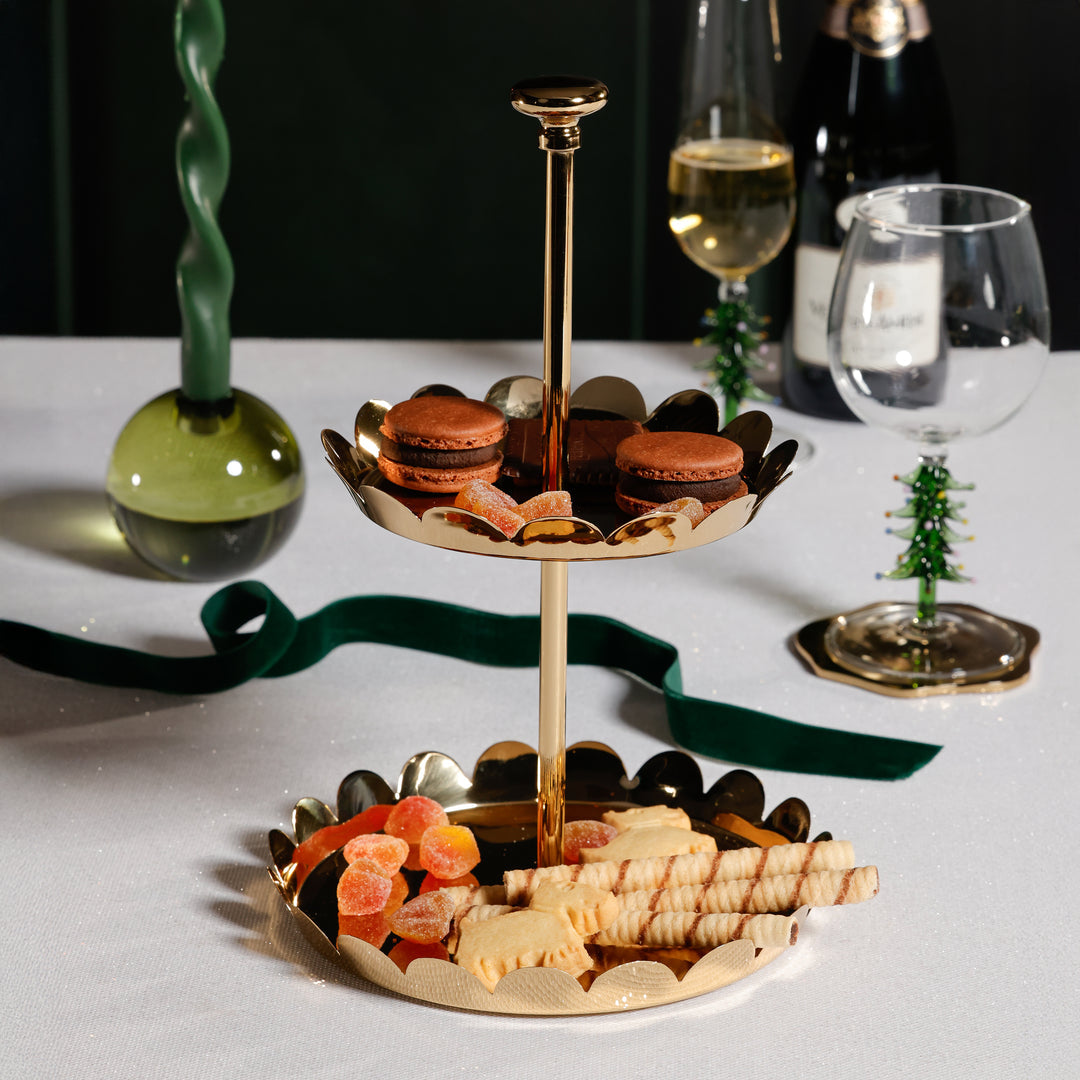 The Zodax Scalloped Two-Tier Tray Collection holds chocolate macarons, heart-shaped cookies, dried fruit, and biscuits on a white tablecloth, surrounded by wine glasses, a green ribbon, and a green candle.