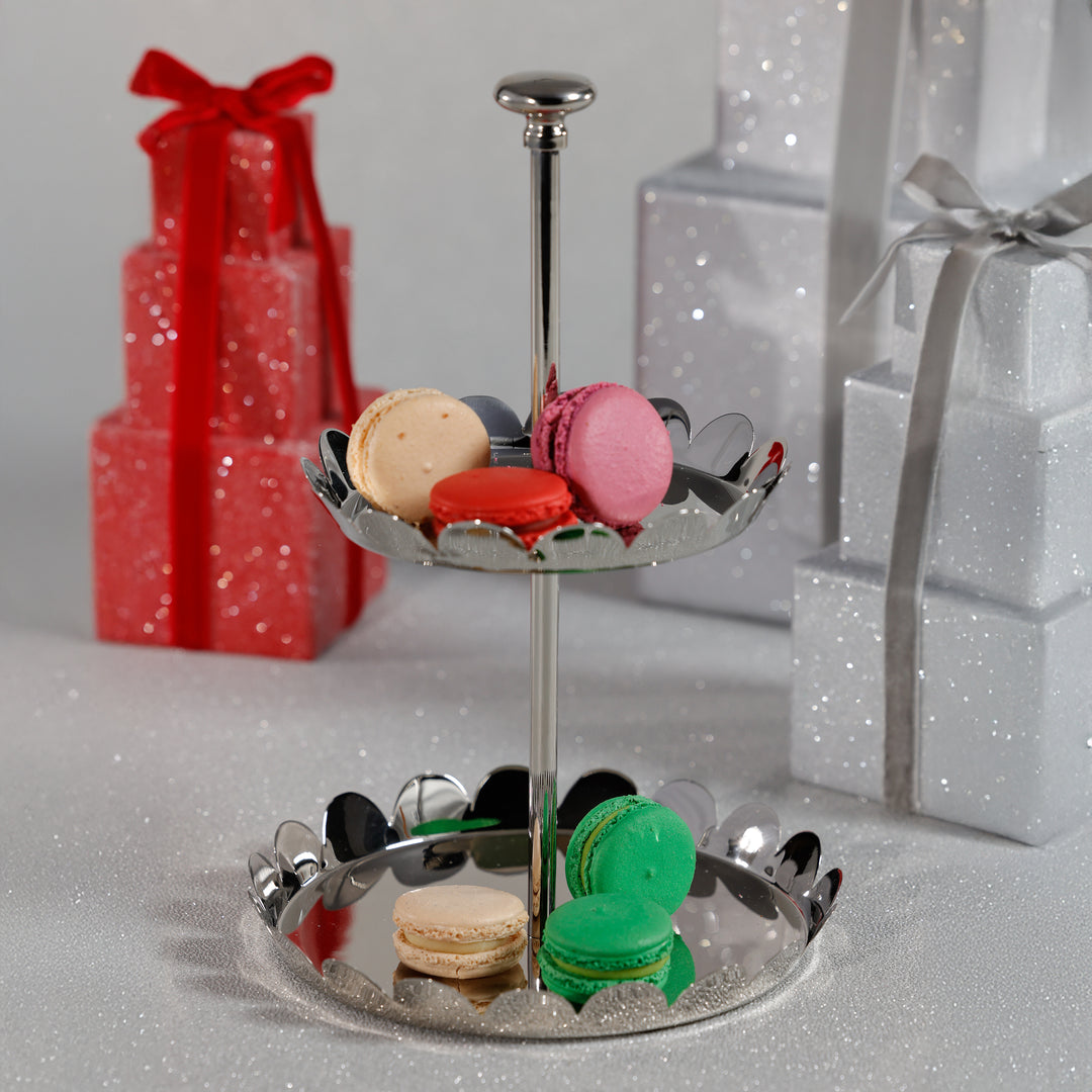 The Zodax Scalloped Two-Tier Tray Collection displays vibrant macarons, while wrapped gift boxes sit behind on a sparkling surface.