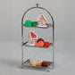 The Zodax High Tea 3-Tier Stand Collection elegantly displays colorful macarons and decorated gingerbread houses, standing out against a plain gray background.