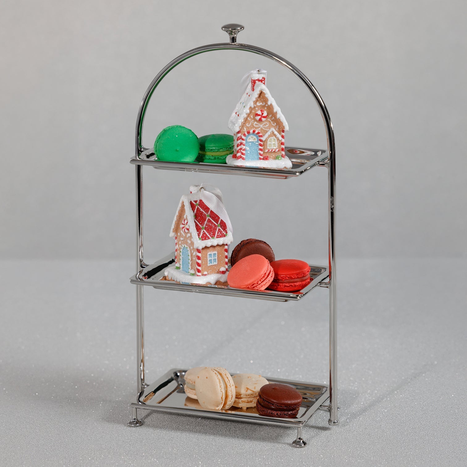 The Zodax High Tea 3-Tier Stand Collection elegantly displays colorful macarons and decorated gingerbread houses, standing out against a plain gray background.