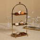 The Zodax High Tea 3-Tier Stand Collection displays assorted macarons and two glass candies for an elegant pastry presentation, set on a beige surface with empty glassware in the background.