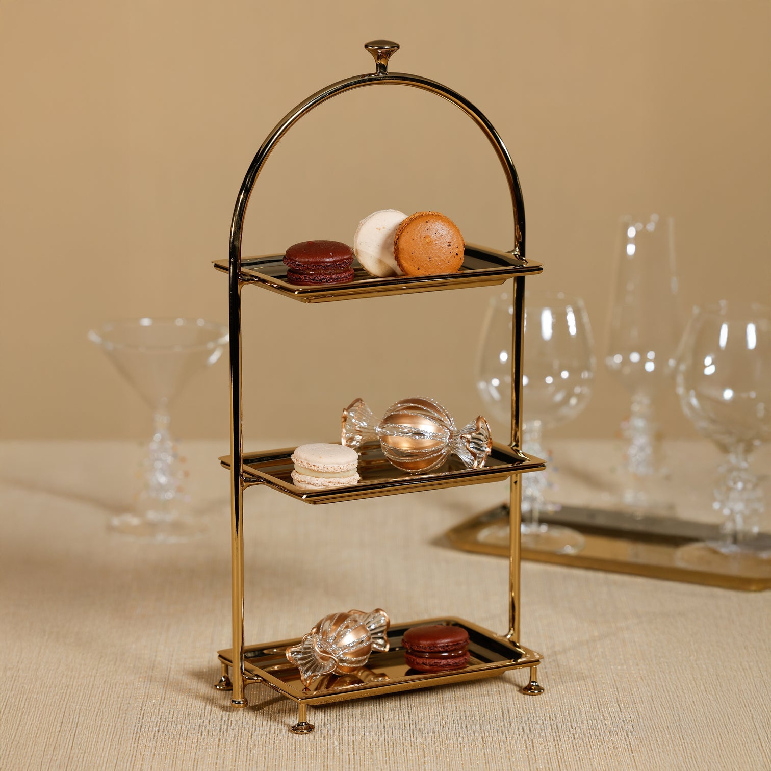 The Zodax High Tea 3-Tier Stand Collection displays assorted macarons and two glass candies for an elegant pastry presentation, set on a beige surface with empty glassware in the background.