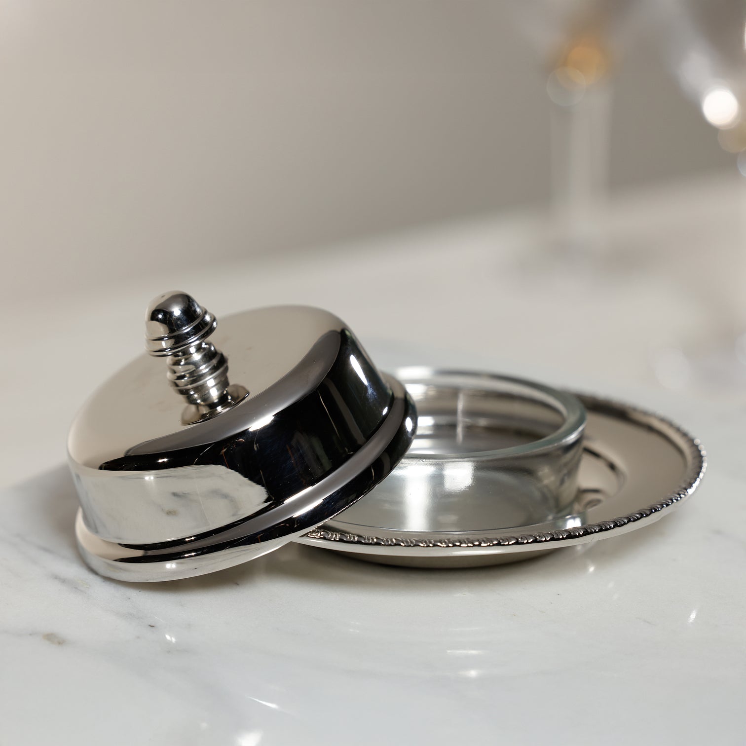 The Zodax Parisian Butter Dish, a small silver piece with a nickel-finished cover and beaded edge detail, rests partially open on a marble surface.