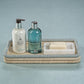 A La Côte Coiled Rectangular Tray by Zodax, woven from abaca fibers, holds Molton Brown body lotion, liquid soap or shower gel, and a bar of soap on a marble dish against a light blue background.