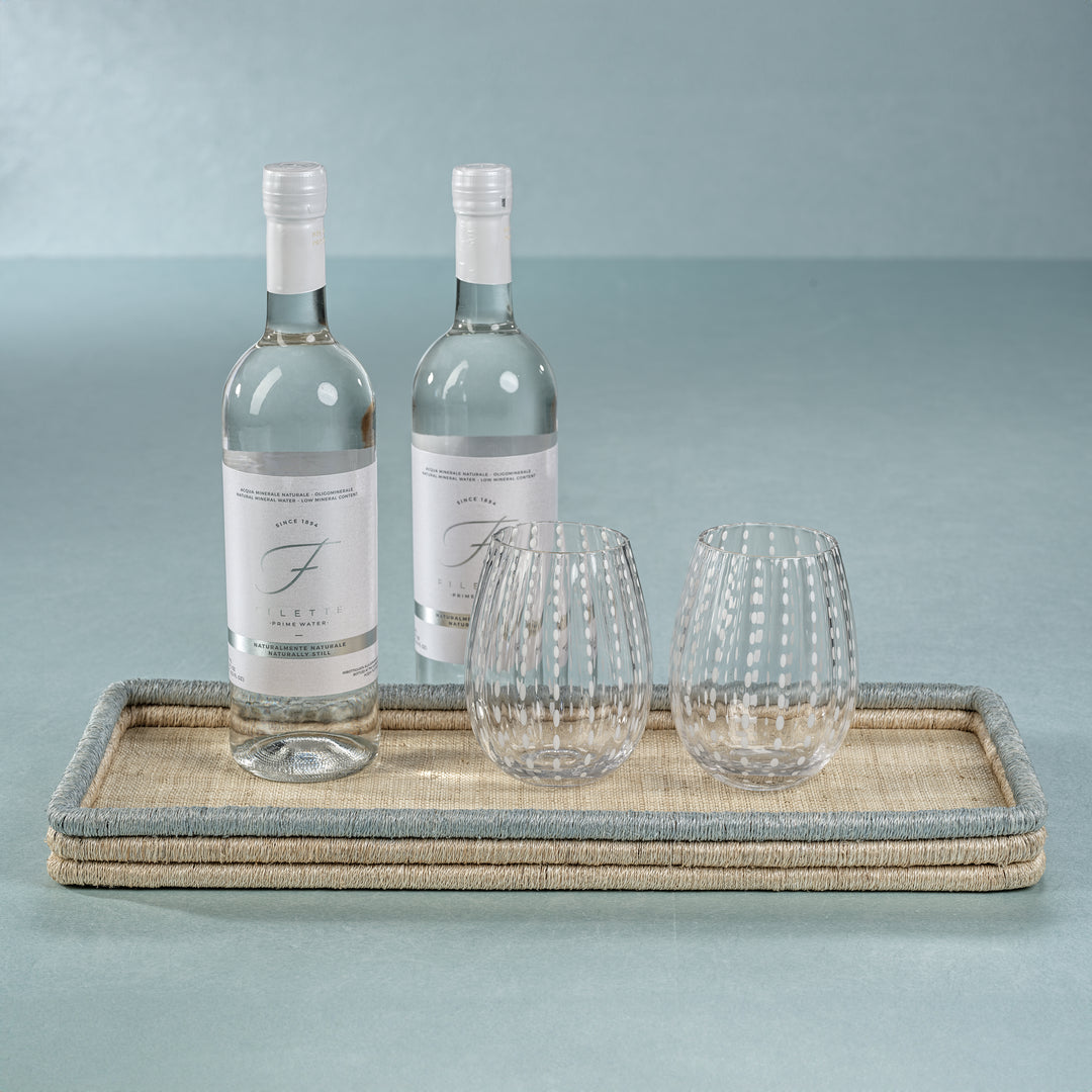Two clear water bottles and two patterned glasses are arranged on a Zodax La Côte Coiled Rectangular Tray, made from abaca fibers, set against a pale blue background.