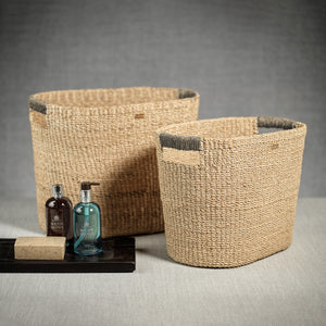 Two Zodax Miramar Oval Abaca Baskets in natural color sit beside a black tray holding two bottles and a bar of soap, all arranged against a neutral backdrop.